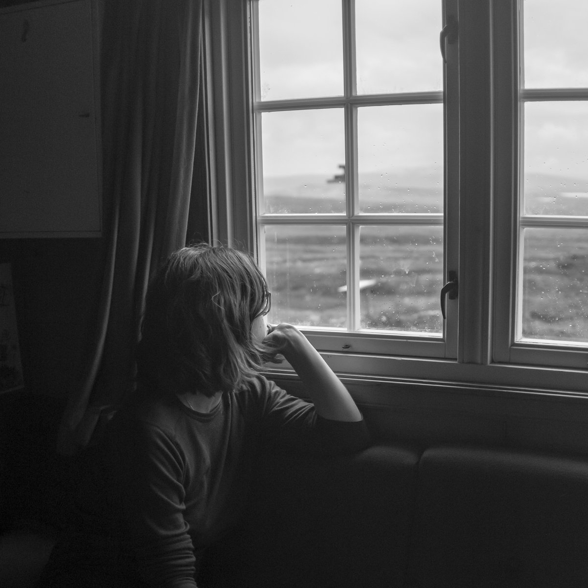 Luisa is looking out of a cabin window onto a blurred, rainy landscape, her back facing the camera and her face hidden behind hair